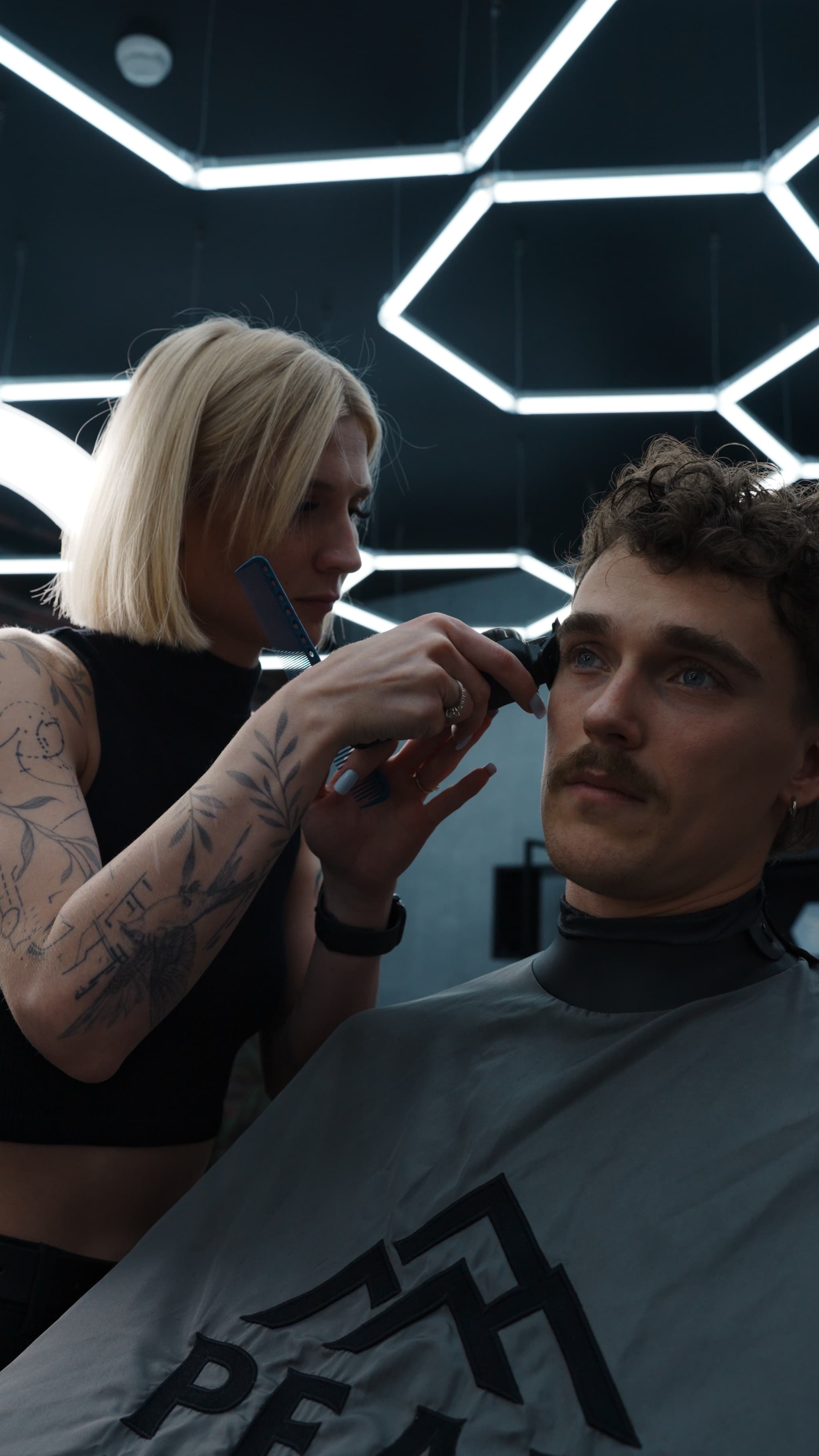 Tetovēta friziere veido vīrieša matu griezumu modernā frizētavā ar ģeometrisku apgaismojumu.